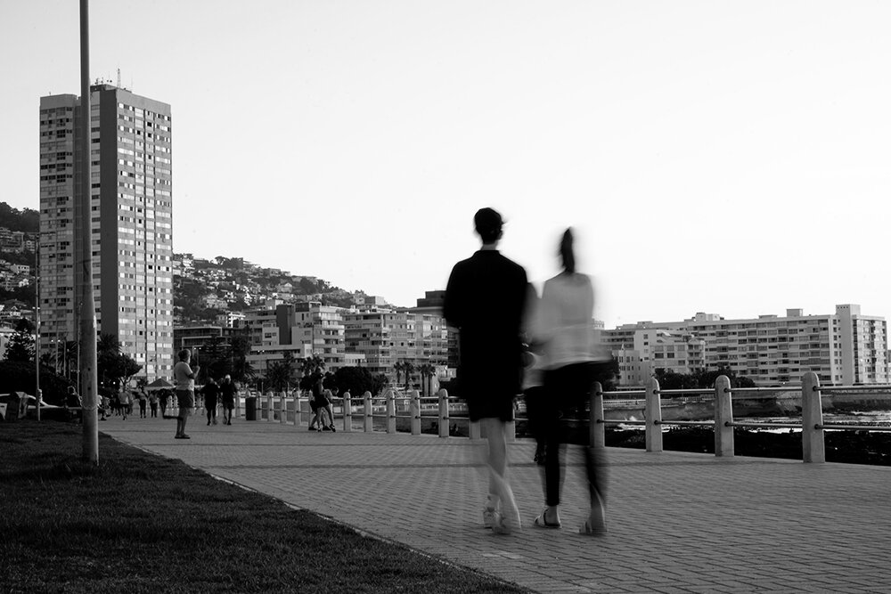 Two people walking on a paved trail near a coast, with tall buildings and more people in the background.