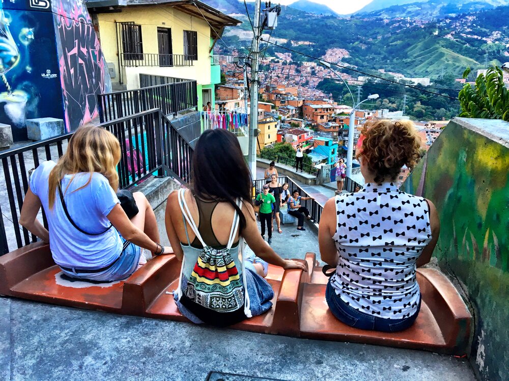 3 women sitting on steps looking out at a colorful view of a town in Colombia