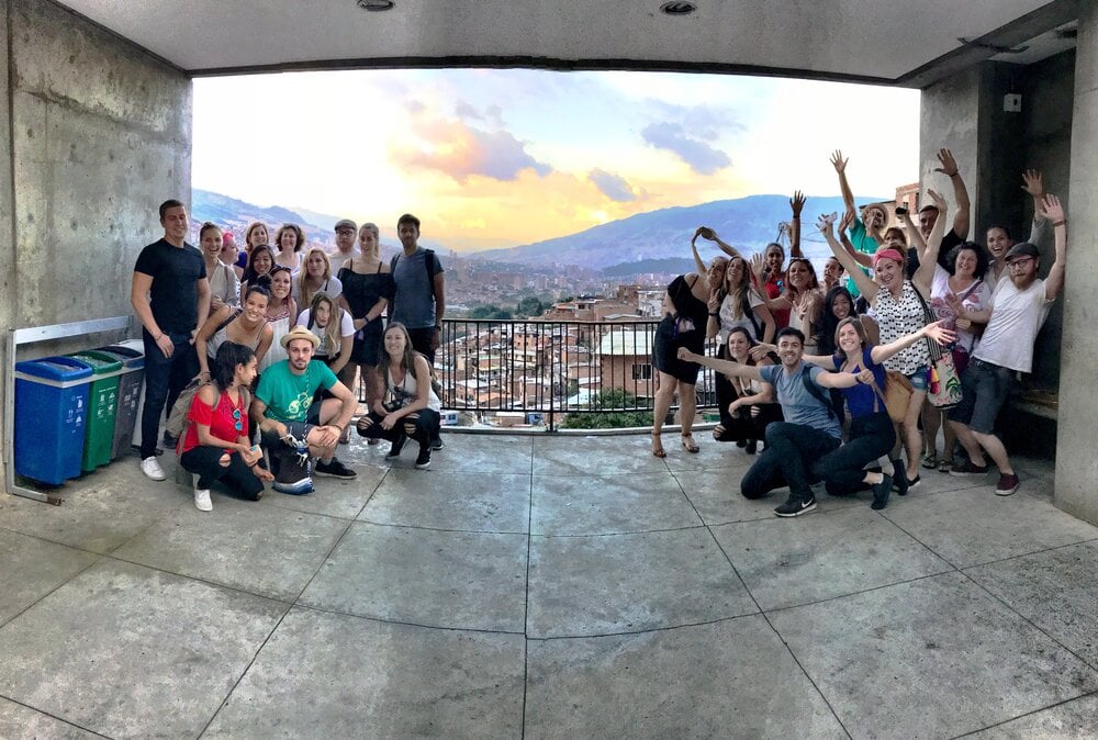 Group of people standing together overlooking a scenic view of a town, mountain range and sunset