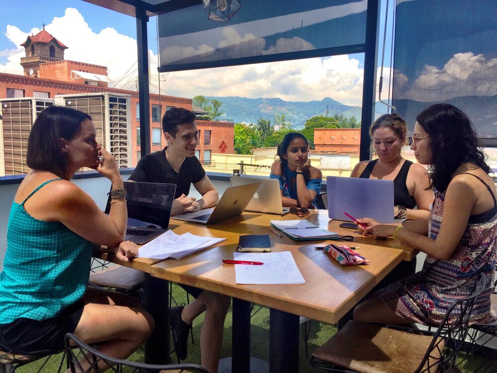 Small group of women and men outside working at a table with laptops and paper with buildings and mountain views in the background